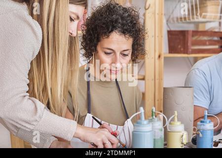Deux potiers féminins se concentrant sur l'application de glaçure à une pièce en céramique dans un atelier de poterie, travaillant en collaboration Banque D'Images