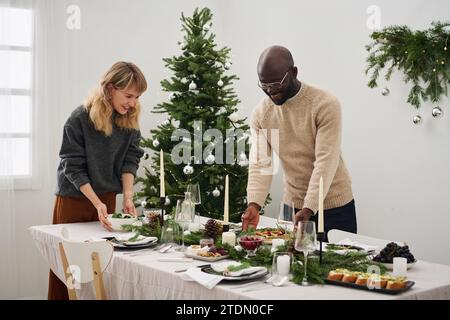 Plan moyen plein de couple biracial plaçant des repas sur la table de noël ensemble se préparant pour la fête Banque D'Images