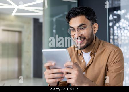 Jeune programmeur hindou près à l'intérieur du bureau en utilisant l'application sur l'ordinateur tablette, développeur masculin testant de nouveaux logiciels, homme d'affaires au lieu de travail portant chemise et lunettes. Banque D'Images