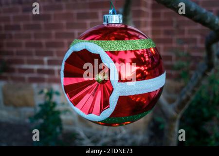 Gros plan d'une boule de Noël rouge vif, blanche et verte suspendue à une branche d'arbre à l'extérieur, un mur de briques flouté à la maison en arrière-plan. Banque D'Images