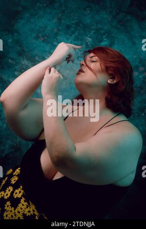 Portrait artistique vertical d'une jeune femme latino Argentine, avec peau blanche, cheveux rouges, taille plus avec jupe florale jaune, debout posant sur cyan b Banque D'Images