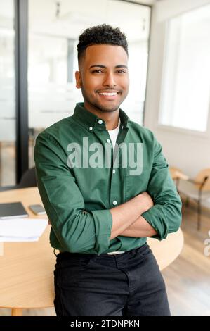 Souriant latin leader hautement qualifié dans les vêtements décontractés regarde la caméra, homme d'affaires hispanique ambitieux confiant se tient au bureau avec les bras pliés, propriétaire d'une startup indienne dans le coworking moderne Banque D'Images