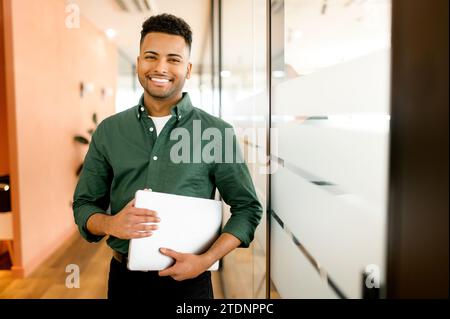 Travailler avec joie. Souriant joyeux homme d'affaires indien portant une chemise verte décontractée debout avec un ordinateur portable dans le bureau, regardant la caméra, fier et ambitieux propriétaire de start-up latine Banque D'Images