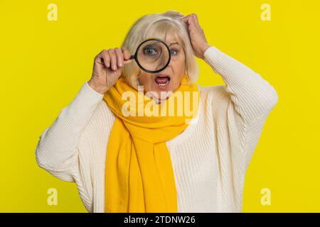 Chercheur chercheur chercheur chercheur senior vieille femme tenant la loupe près du visage, regardant dans la caméra avec grand oeil drôle zoomé, recherchant, analysant. Grand-mère âgée sur fond jaune Banque D'Images