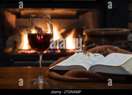 Verre de vin rouge avec couverture et livre sur la table en bois. Cheminée en feu dans bqckground Banque D'Images