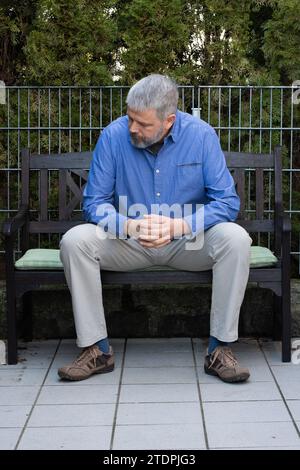 Homme aux cheveux gris et à la barbe pleine, 50-59 ans, assis sur un banc sur la terrasse à l'extérieur, pendant la tête, réfléchi, triste Banque D'Images