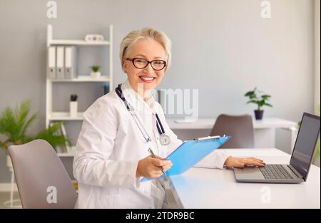 Le médecin expérimenté aux cheveux gris souriant se tourne vers la caméra après avoir consulté le patient via un ordinateur portable Banque D'Images