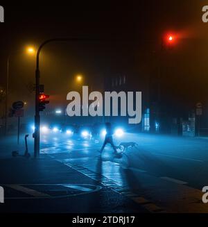 Une personne traverse la route avec un chien sur une nuit brumeuse avec la lumière flash de voiture et l'éclairage de rue Banque D'Images