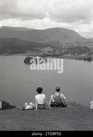 Années 1950, historique, un homme et une femme dehors marchant, ayant une pause assis sur un terrain plus élevé, regardant sur le lac et le paysage environnant, Yorkshire, Angleterre, Royaume-Uni, l'homme avec sac à dos en toile sur son dos. Banque D'Images