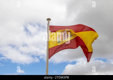 Un grand fragment rouge-jaune de l'Espagne sur un mât de drapeau se développe dans un vent fort sur un fond de ciel bleu et de nuages blancs Banque D'Images