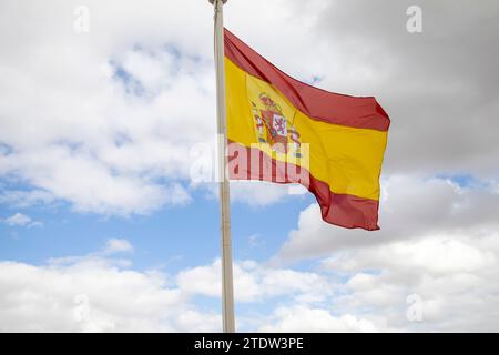 Un grand fragment rouge-jaune de l'Espagne sur un mât de drapeau se développe dans un vent fort sur un fond de ciel bleu et de nuages blancs Banque D'Images