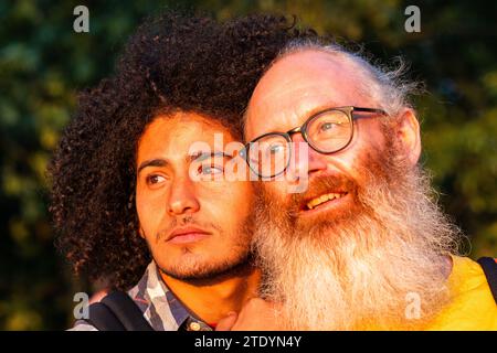 Dans cette image réconfortante, deux individus masculins, peut-être gays, partagent un moment de l’heure d’or. Leurs regards sont dirigés au loin, suggérant la contemplation ou une vision partagée. L'homme plus âgé, avec sa barbe blanche distinctive et ses lunettes, sourit largement, exsudant un sentiment de joie et de sagesse. Les cheveux bouclés et l'expression sincère de la jeune personne véhiculent un comportement réfléchi. La lumière chaude baigne leurs visages, soulignant les contrastes et les similitudes entre eux et créant une atmosphère de connexion intime. Compagnie Golden Hour. Photo de haute qualité Banque D'Images