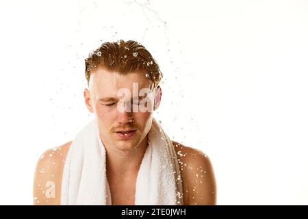 Routine matinale. Heureux, beau modèle masculin roux lavé visage dans les éclaboussures d'eau sur fond de studio blanc. Banque D'Images