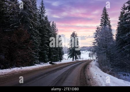 route asphaltée à travers la campagne boisée en hiver au coucher du soleil. arbres et collines dans la neige. nuages dramatiques sur le ciel Banque D'Images