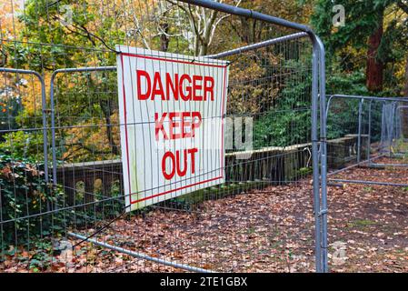 Un grand panneau d'avertissement de danger de rester à l'extérieur monté sur une clôture temporaire dans un parc public avec une maçonnerie de parapet instable derrière Chertsey Surrey Angleterre Royaume-Uni Banque D'Images