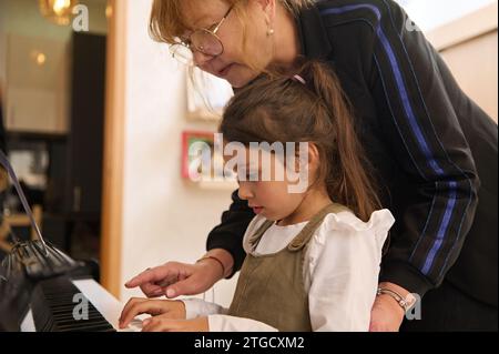 Gros plan enfant caucasien d'âge élémentaire, une charmante petite étudiante assise au piano tandis qu'une professeure de pianiste l'enseigne, donnant de la musique lesso Banque D'Images