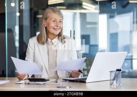 Comptable féminin senior financier au lieu de travail à l'intérieur de la paperasse de bureau, femme d'affaires satisfaite des résultats de réalisation des rapports de vérification, des contrats patron tenant le papier dans les mains, ordinateur portable de travail. Banque D'Images