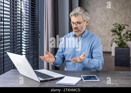 Homme d'affaires concentré sérieux parlant à l'intérieur du bureau assis à table avec ordinateur portable, patron aux cheveux gris senior utilisant nuotbook pour la communication à distance réunion en ligne avec des collègues partenaires. Banque D'Images