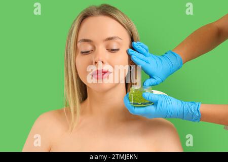 Esthéticienne appliquant un gommage sur le visage de jeune femme sur fond vert Banque D'Images