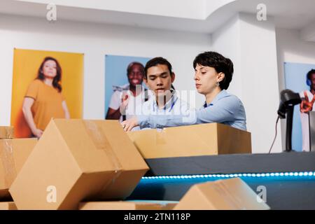 Divers employés travaillant aux commandes des clients, préparant des colis pour la livraison dans une boutique moderne. Les travailleurs mettant des vêtements à la mode dans des boîtes en carton, discutant de detalis d'expédition au comptoir Banque D'Images