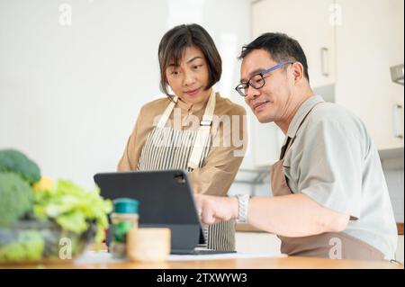 Les couples asiatiques adultes charmants et heureux regardent une recette en ligne sur une tablette numérique et cuisinent dans la cuisine ensemble. liens familiaux, cuisine maison Banque D'Images