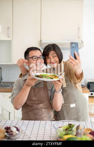 Un charmant couple asiatique prenant des photos ou selfies avec leur nourriture dans la cuisine ensemble. vie domestique, liens familiaux, mari et femme Banque D'Images