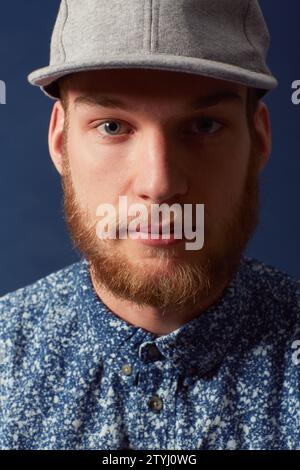 Homme, mode et visage avec chapeau dans un studio, denim et tendance isolé sur fond bleu. Modèle irlandais avec barbe hipster, gingembre et sérieux Banque D'Images