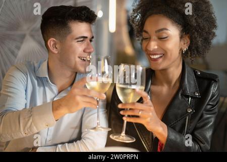 Un jeune couple partage un toast avec des verres de vin blanc, profitant d'un moment intime ensemble dans un cadre chaleureux de restaurant. Banque D'Images