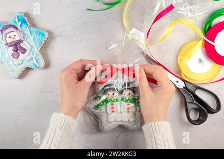 les mains féminines dans un pull blanc attachent un arc rouge sur un paquet de biscuits en pain d'épice avec des bonhommes de neige sur le fond d'une table lumineuse, des rubans multicolores et des ciseaux. Concept pour les cadeaux de Noël ou du nouvel an faits soi-même. Banque D'Images