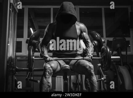 Image en noir et blanc d'un athlète puissant dans un sweat à capuche assis sur un banc dans une salle de gym. Concept de fitness et de musculation Banque D'Images