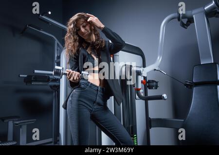 Belle grande femme aux cheveux bruns dans un costume posant dans la salle de gym avec un haltère. Sport et concept d'affaires. Supports mixtes Banque D'Images