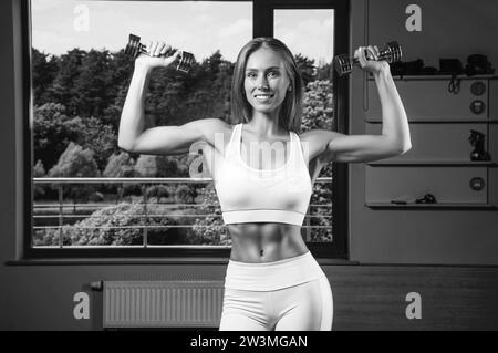 Charmant jeune athlète s'entraîne avec des haltères dans la salle de gym. Concept de culturisme. Sport. Haltérophilie. Supports mixtes Banque D'Images