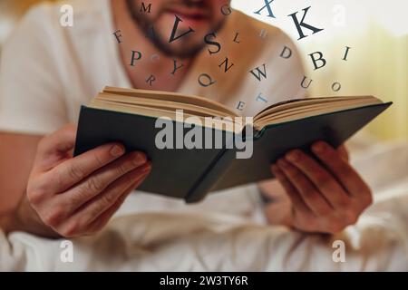 Jeune homme lisant le livre avec des lettres volant au-dessus sur le lit à la maison, closeup Banque D'Images