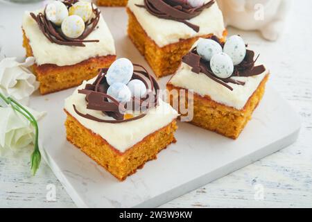 Barres de gâteau aux carottes de Pâques décorées avec nid de chocolat et œufs de bonbons au chocolat fleurissant des fleurs de cerise ou de pomme sur des fonds rustiques en bois clair. Banque D'Images