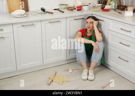 Femme fatiguée assise sur le sol sale avec des ustensiles et des restes de nourriture dans la cuisine désordonnée Banque D'Images