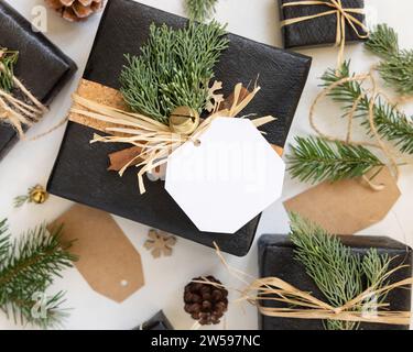 Cadeau noir avec étiquette cadeau de Noël, branches de sapin, pommes de pin et décorations de vacances, vue de dessus. Composition hivernale avec étiquette vierge maquette, co Banque D'Images