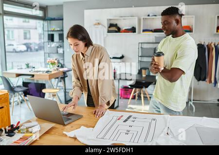 homme afro-américain avec du café pour aller près de collègue asiatique travaillant sur ordinateur portable dans le studio d'impression Banque D'Images