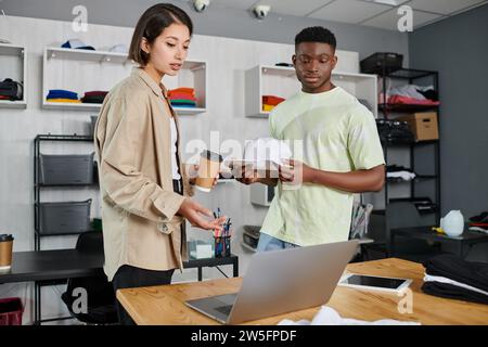 designers multiethniques avec casquette blanche et tasse de papier regardant ordinateur portable sur le bureau de travail dans le studio d'impression Banque D'Images