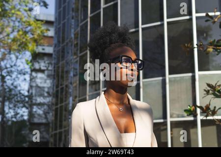 Portrait de souriant noir élégant jeune femme entrepreneure avec des cheveux bouclés noir chignon portant des lunettes et des formals tout en se tenant contre le bâtiment i. Banque D'Images