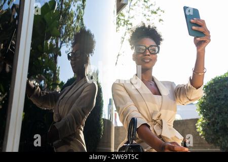 Angle bas de belle jeune femme noire Manager dans les formals et lunettes avec rouge à lèvres assis par le mur de verre et regardant smartphone tout en prenant Banque D'Images