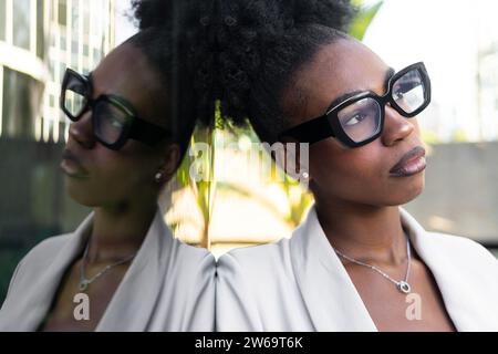 Gros plan de belle jeune femme d'affaires afro-américaine avec les cheveux noirs et les lunettes contemplant tout en s'appuyant sur le mur de verre Banque D'Images