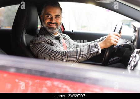 Heureux homme d'affaires indien barbu assis dans l'automobile et regardant loin par la fenêtre ouverte tout en conduisant dans la rue Banque D'Images