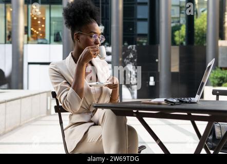 Professionnel féminin afro-américain concentré avec des lunettes et des cheveux noirs tenant la tasse de café et regardant l'ordinateur portable tout en travaillant au café de trottoir Banque D'Images