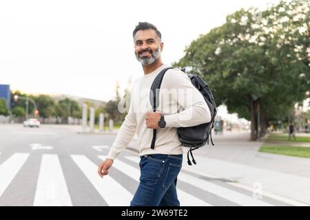 Vue latérale de souriant Bearded homme d'affaires adulte portant un chandail blanc avec sac à dos traversant la rue et regardant loin contre les arbres de fond Banque D'Images