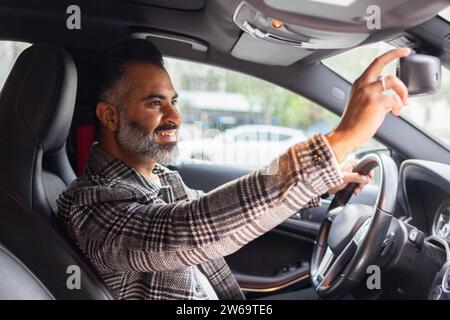 Vue latérale du conducteur indien assis dans la voiture et regardant loin avec plaisir tout en ajustant le rétroviseur dans la journée Banque D'Images