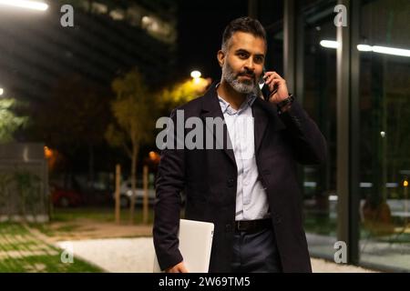 Entrepreneur indien concentré dans la tenue chic tenant l'ordinateur portable et parlant sur smartphone après réunion d'affaires réussie Banque D'Images