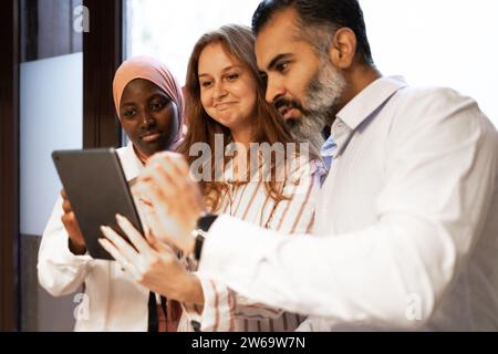 Positif divers collègues portant des vêtements décontractés debout tout en regardant une tablette tout en ayant une pause-café dans un bureau moderne Banque D'Images