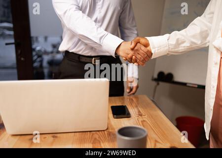 Crop homme d'affaires anonyme se serrant la main et saluant les uns les autres tout en se tenant près de l'ordinateur portable avec téléphone portable sur la table en bois dans la salle de bureau moderne ag Banque D'Images