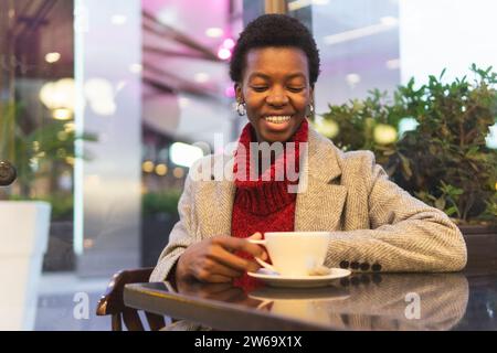 Souriante femme afro-américaine en manteau assise à table près du mur de verre buvant une boisson chaude pendant le week-end Banque D'Images