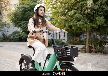 Une jeune femme portant un casque monte un vélo électrique vert dans une rue de la ville, favorisant le transport urbain écologique. Banque D'Images